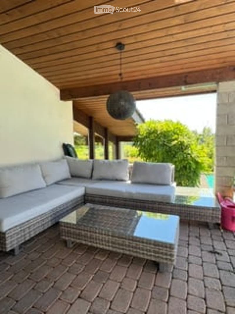 Outdoor patio with wooden ceiling, grey sectional sofa, glass coffee table, brick floor, and swimming pool visible in the background.