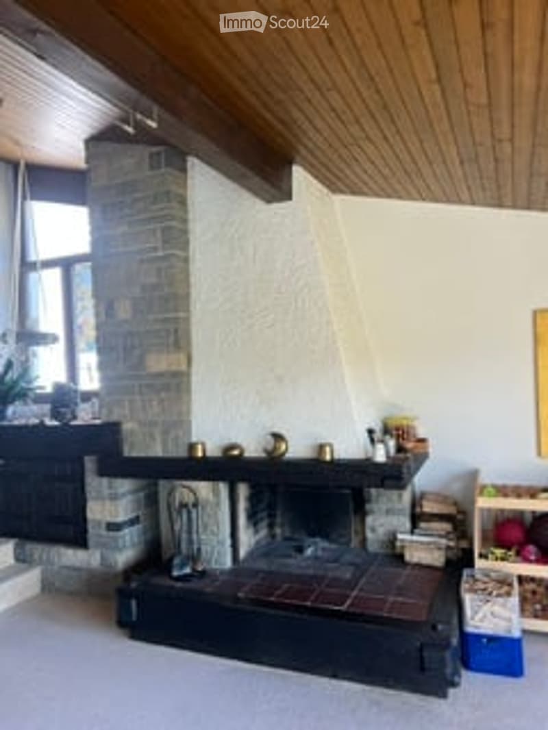 Cozy living room with a stone fireplace, wooden ceiling beams, and a large window. A small table and chair are positioned near the fireplace, along with decorative items like vases and a picture frame on the wall.