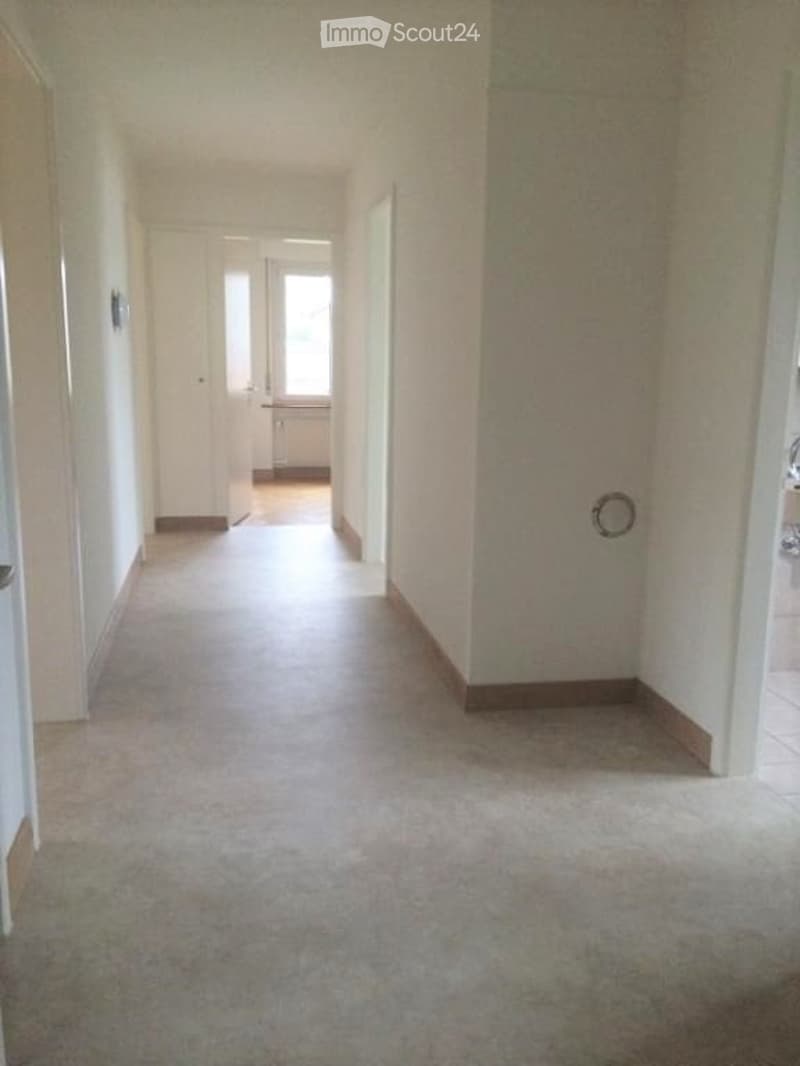 Empty hallway with white walls and flooring, doors on both sides, and a window at the end.