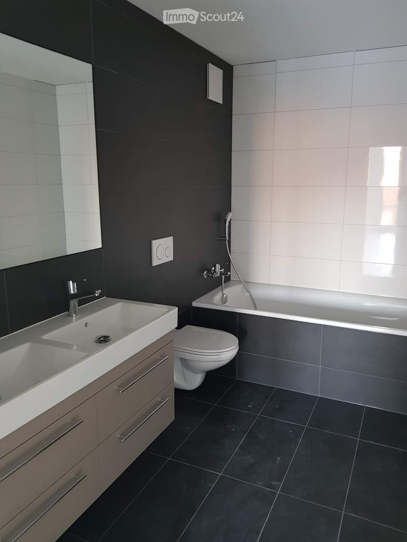 Modern bathroom with dark grey tiles, white sink, toilet, bathtub, and mirror. Features a sleek, contemporary design with a large window providing natural light.