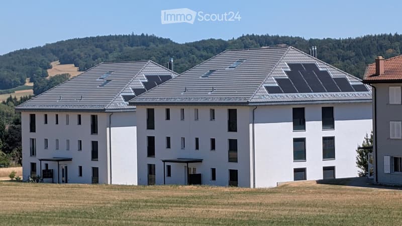 Modern two-story building with white walls, grey roof, multiple windows, and solar panels on the roof, set in a grassy field with a forested hill in the background.