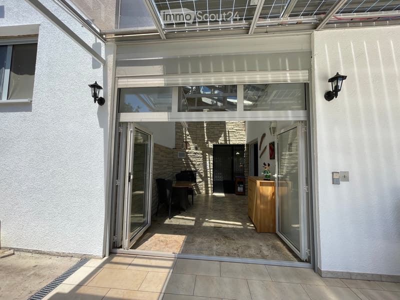 A spacious, well-lit balcony or terrace area with large glass doors leading into a room. The interior features a dining table, chairs, and a wooden cabinet. The floor is tiled, and the walls are white.