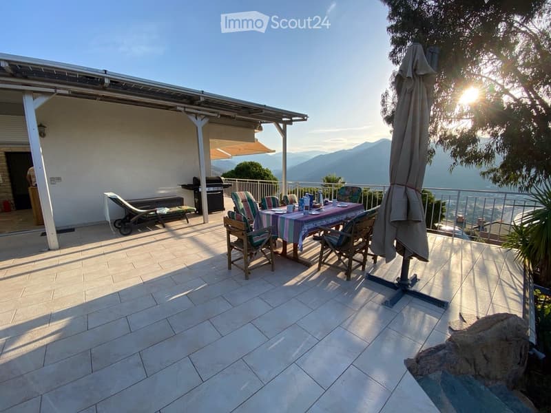Balcony with tiled floor, outdoor dining table and chairs, umbrella, sun loungers, and a grill, overlooking a scenic mountain view.