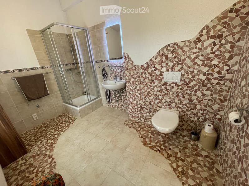 Bathroom with a toilet, sink, and glass-enclosed shower. Features mosaic tile accents on walls and floor, a towel on a rack, and a toilet paper holder.