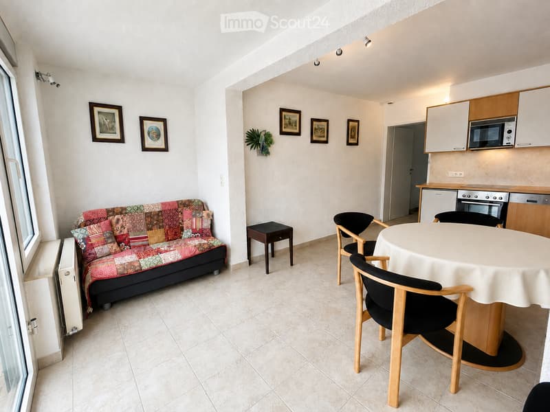 A compact, well-furnished living and dining area features a colorful patchwork sofa, a small wooden stool, a round dining table with four chairs, and a modern kitchen area with cabinets, a microwave, oven, and dishwasher. The space is decorated with frame
