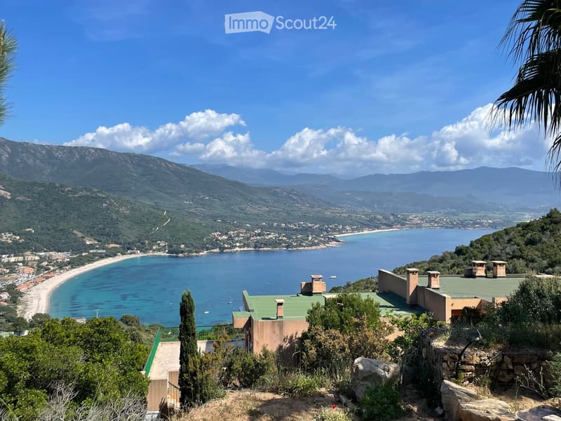 A scenic coastal view from a hillside property, featuring a house with a green roof, surrounded by lush greenery, overlooking a sandy beach and the Mediterranean Sea with distant mountains.