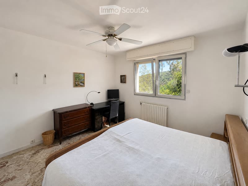 Cozy bedroom with a wooden bed, desk with a computer, chest of drawers, and a view of greenery through large windows.