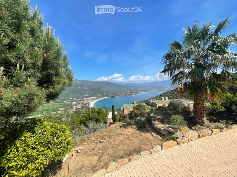 A scenic outdoor view from a hillside, featuring a brick pathway, lush greenery, a palm tree, and a distant coastal town with a beach and mountains in the background.