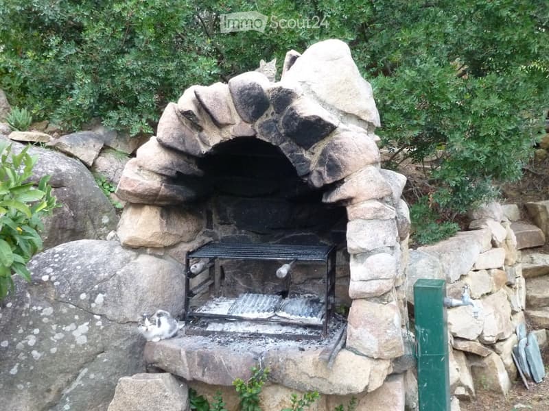 Stone outdoor fireplace with a metal grill, surrounded by rocks and greenery, with two cats nearby.