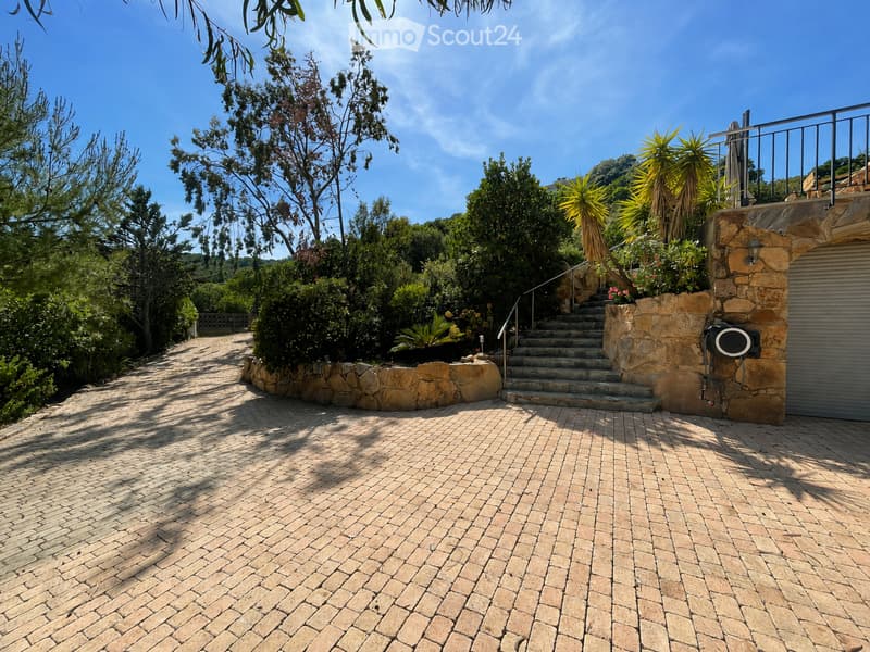 Brick-paved outdoor area with stone walls, steps leading up, lush greenery, and a clear blue sky.