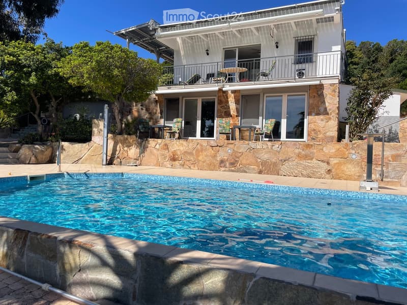 A modern two-story house with a swimming pool in the foreground, surrounded by a stone wall and outdoor furniture on the patio and balcony.