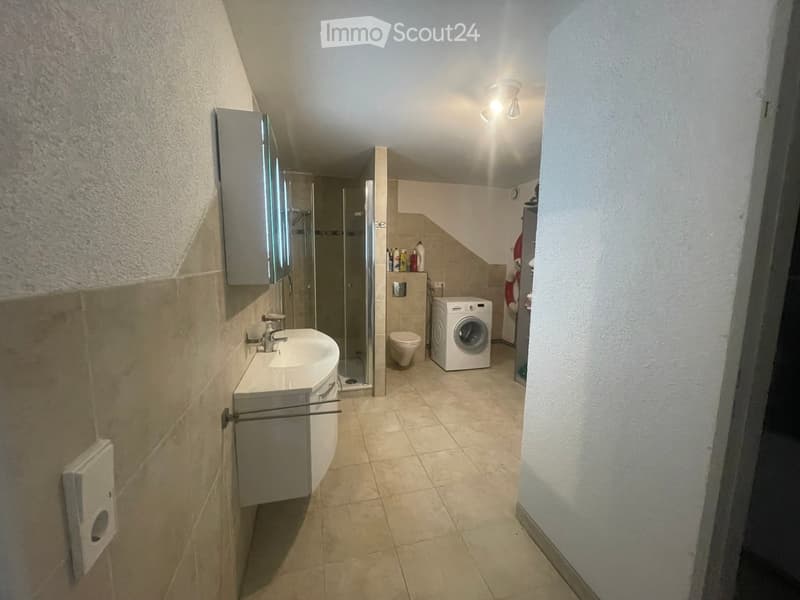 Modern bathroom with a sink, mirror, toilet, washing machine, and a glass-enclosed shower. The space features beige tiled floors and walls, with a small shelf holding toiletries.