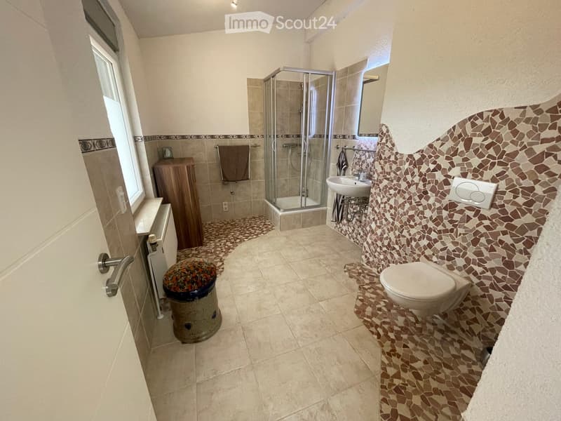 Modern bathroom with beige tiles, a glass-enclosed shower, a toilet, a sink, a wooden cabinet, and a decorative stool.