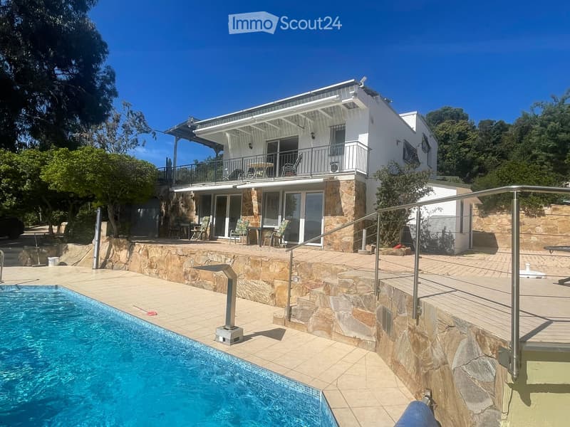 Modern two-story house with a swimming pool, stone terraces, and outdoor seating areas. Features include a balcony on the upper level and a staircase leading to the pool area.