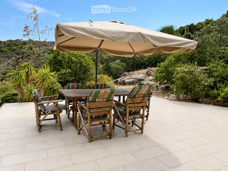 An outdoor patio area with a large umbrella, wooden table, and several wooden chairs with striped cushions, surrounded by lush greenery and a stone pathway.