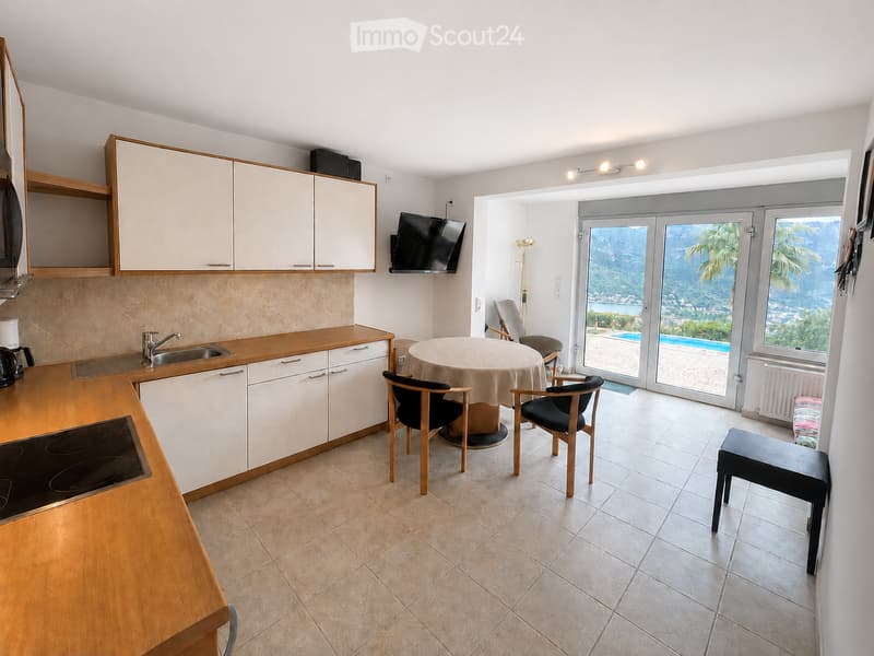 A modern kitchen and dining area with white cabinets, a wooden countertop, an electric stove, a sink, and a coffee maker. A round dining table with chairs is placed near a large sliding glass door that opens to a balcony with a view of the sea and mountai