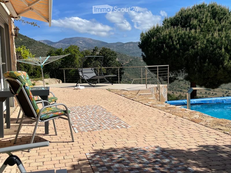Outdoor patio with brick tiles, lounge chairs, a swing chair, a small table, and a swimming pool, overlooking a scenic mountain view.