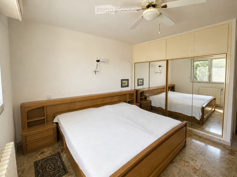 Spacious bedroom with a large wooden bed, mirrored wardrobe, built-in cabinets, and a ceiling fan. The room features tiled flooring and a small patterned rug.