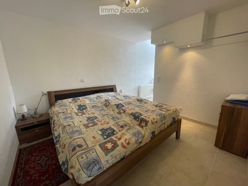 A furnished bedroom with a wooden bed, patterned bedspread, nightstand with lamp, and a wooden drawer unit.