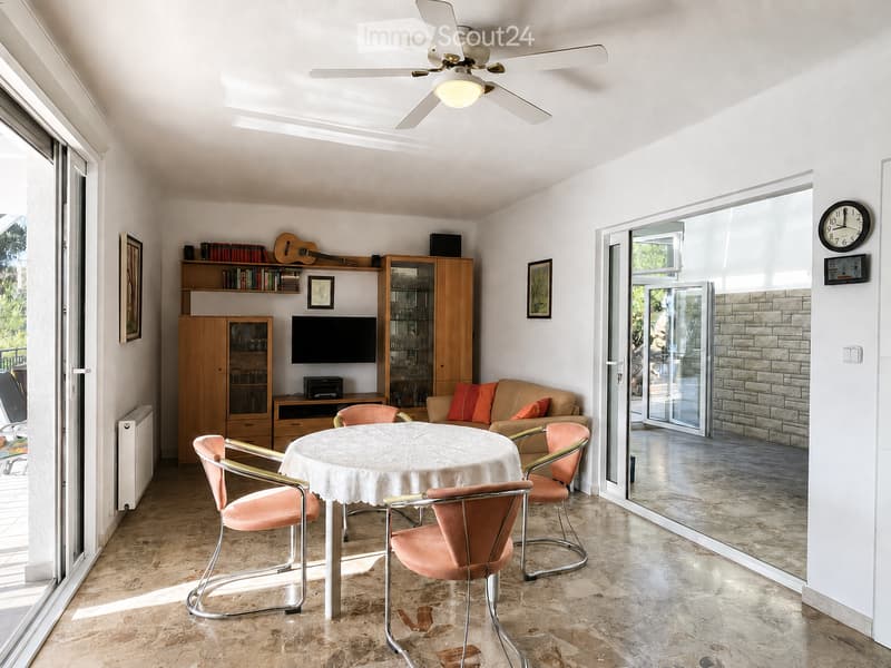Bright living room with a round table, four chairs, a sofa with orange cushions, a wooden cabinet, a guitar, and a television. Large sliding glass doors lead to a balcony.