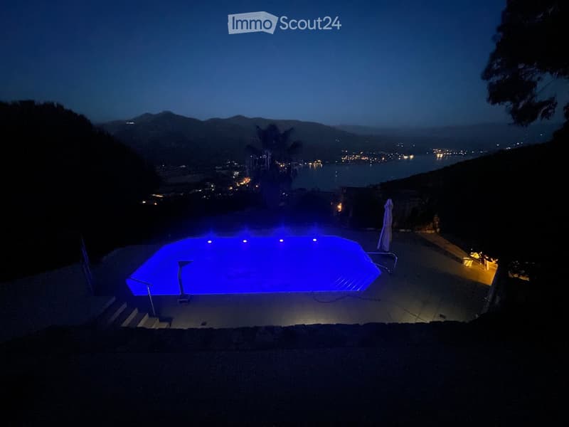 Nighttime view of a swimming pool with blue lighting, overlooking a coastal town and water with distant lights.