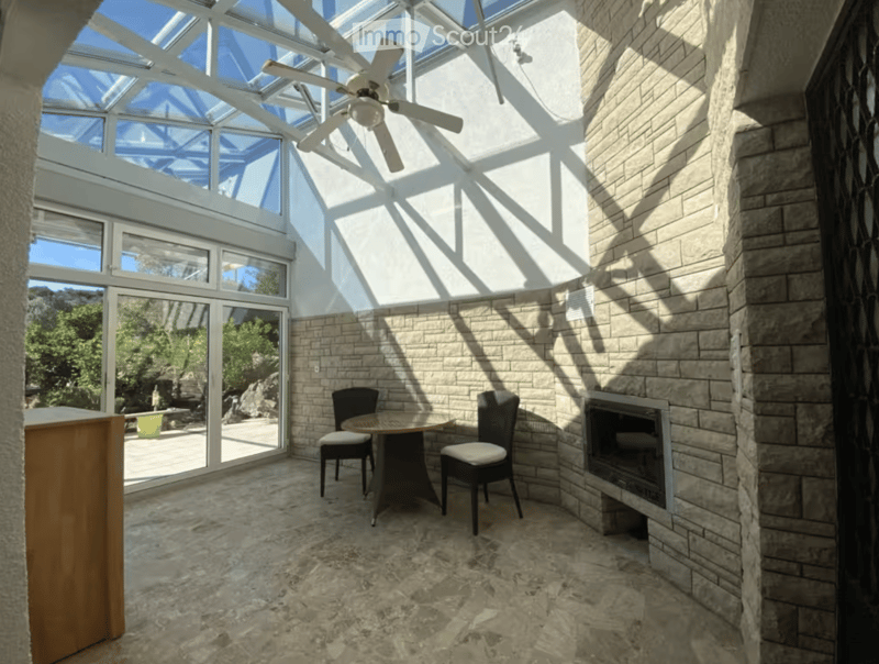 Bright sunroom with a stone fireplace, dining table, chairs, and ceiling fan. Large windows and glass doors open to outdoor space.