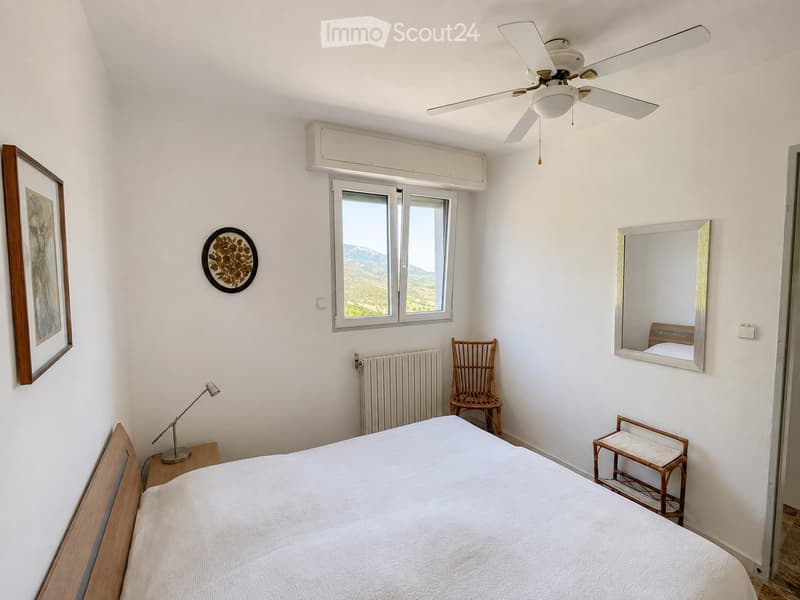 Bright bedroom with a white bed, wooden nightstand, lamp, wicker chair, mirror, and a scenic mountain view through a large window.