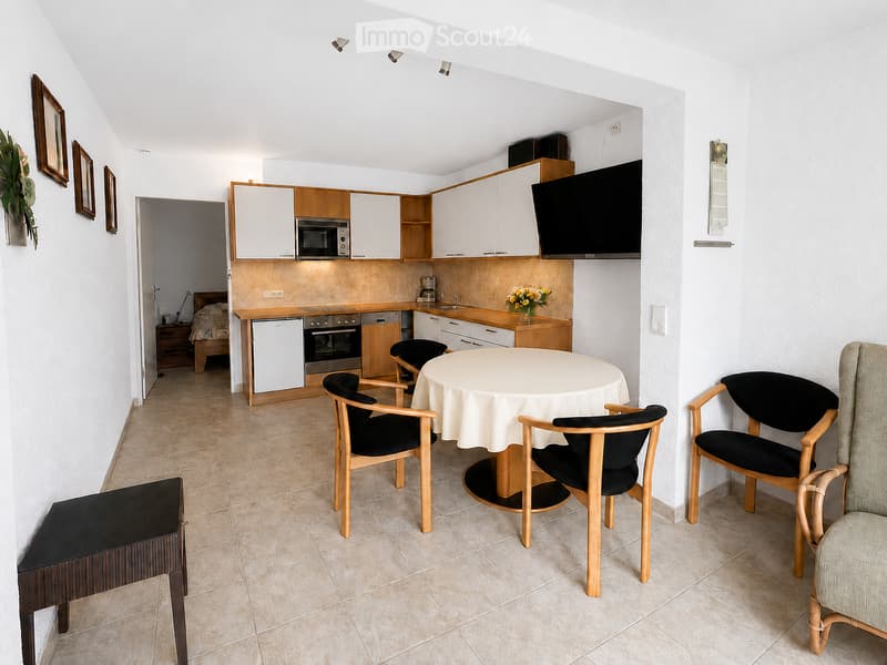 A compact, well-furnished living and dining area with a small kitchen. The space includes a round dining table with four chairs, a television mounted on the wall, and a small kitchen area with cabinets, a microwave, oven, dishwasher, and refrigerator. The