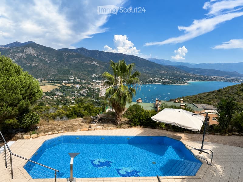 Outdoor area with a swimming pool, surrounded by a tiled patio, featuring a large umbrella, palm trees, and a scenic mountain and sea view.