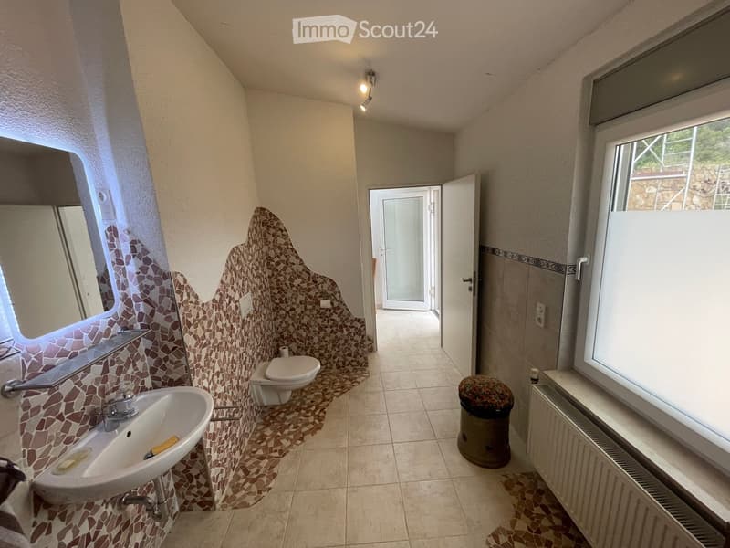 Bathroom with mosaic tile accents, a sink, toilet, mirror, radiator, and a trash can. The room has large windows and an open door leading to another space.