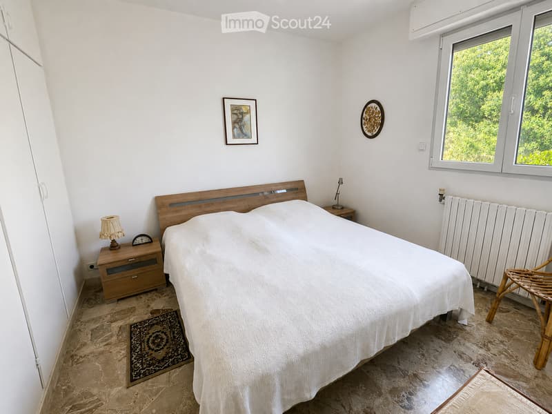 Cozy bedroom with a large bed, wooden nightstand, lamp, and a wicker chair near the window. The room features a patterned rug and a view of greenery outside.