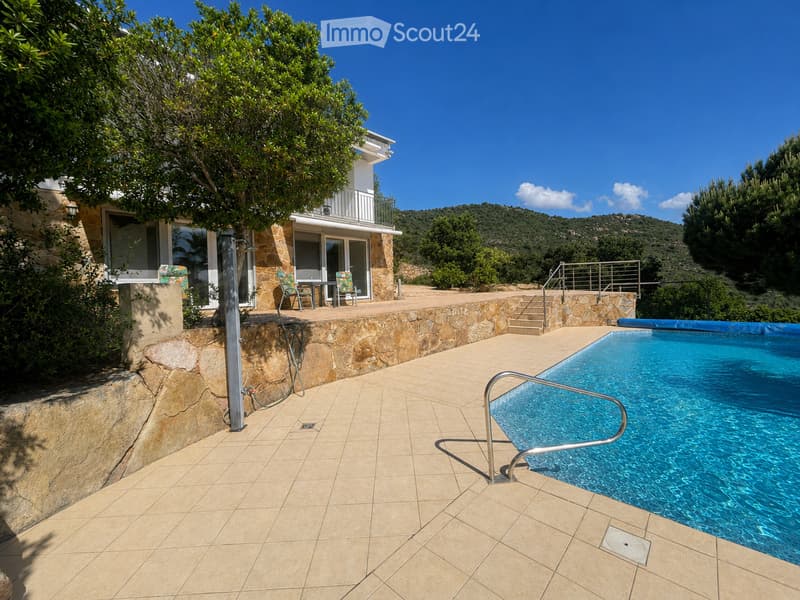 Outdoor area with a swimming pool, tiled patio, stone wall, and a modern house with a balcony and large windows in the background.
