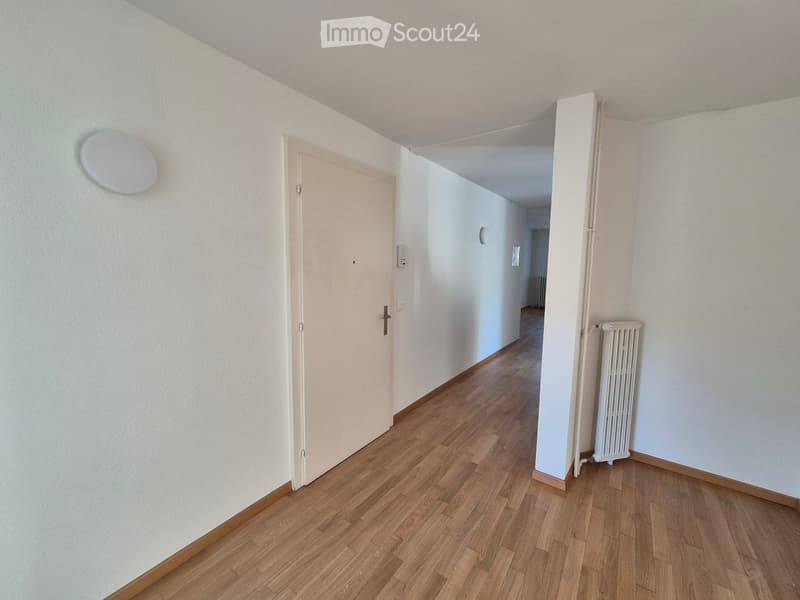 Empty hallway with wooden flooring, white walls, and a closed door on the left. A radiator is visible on the right wall, and a light fixture is mounted on the left wall.