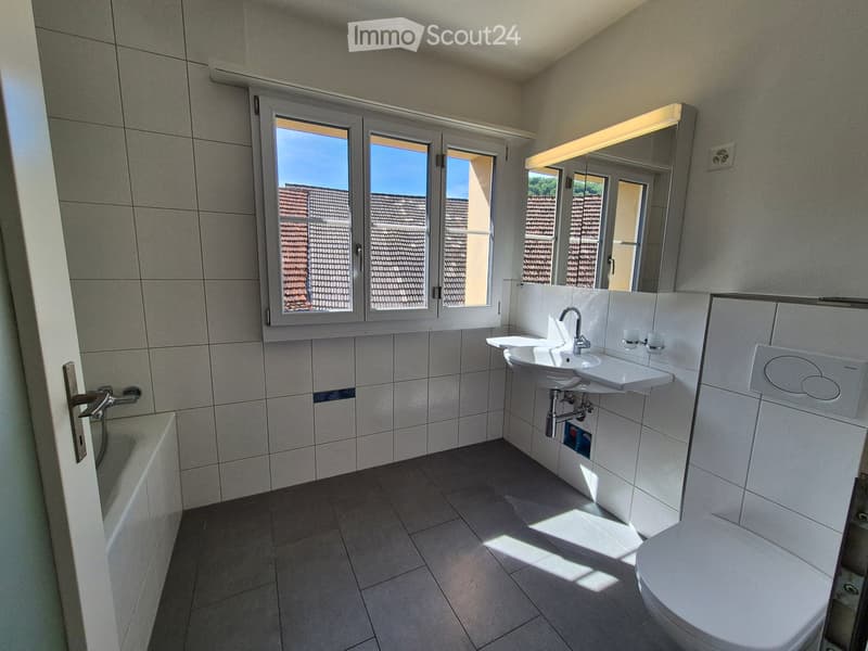 Modern bathroom with tiled walls and floor, featuring a bathtub, sink with faucet, toilet, and a large mirror above the sink. Natural light streams in through a window with white frames.