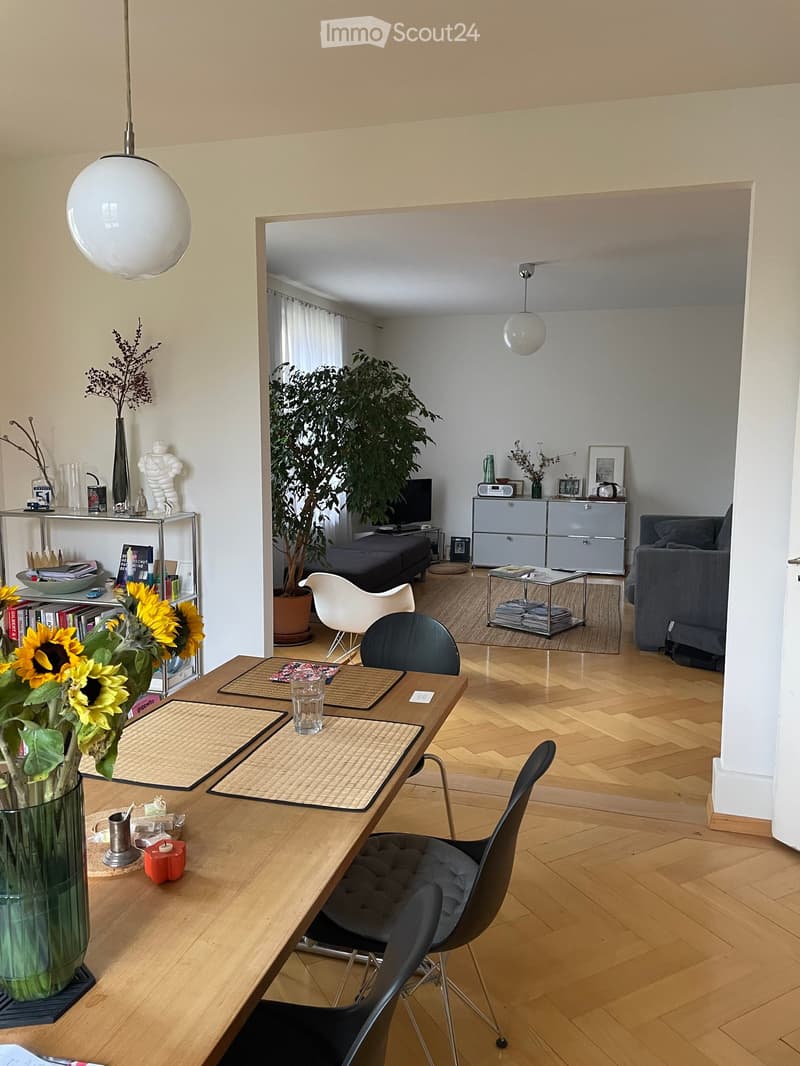 The image shows a modern, well-lit living room with a wooden floor, a large round pendant light, and a dining area with a wooden table and chairs. There is a sofa, a cabinet, and a plant in the background.