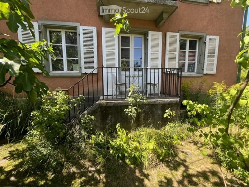 Two-story building with a balcony, white shutters, and a small garden area.