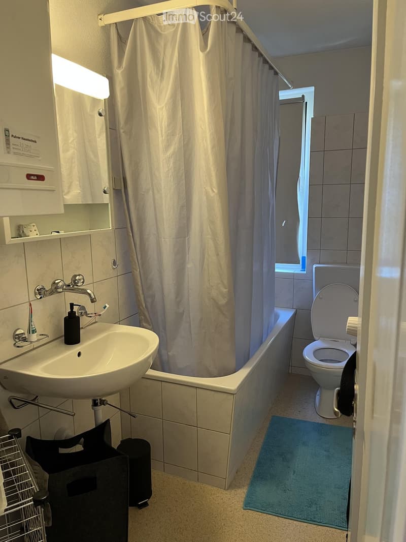 Modern bathroom with a white sink, bathtub with a shower curtain, toilet, and a small blue rug on the floor. The sink has a faucet, toothbrush, toothpaste, and a soap dispenser. A mirror is mounted above the sink, and a small cabinet is visible on the lef