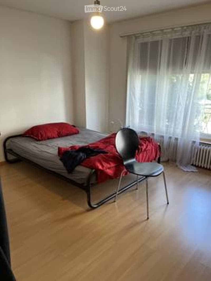 A small, furnished bedroom with a single bed, a chair, and a pillow. The room features wooden flooring and large windows with sheer curtains.