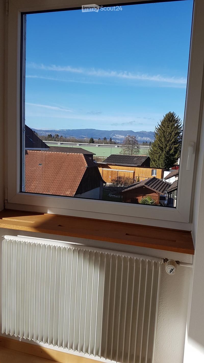 View from a window showing a clear blue sky, distant hills, and neighboring houses with red-tiled roofs. A white radiator is visible below the window.