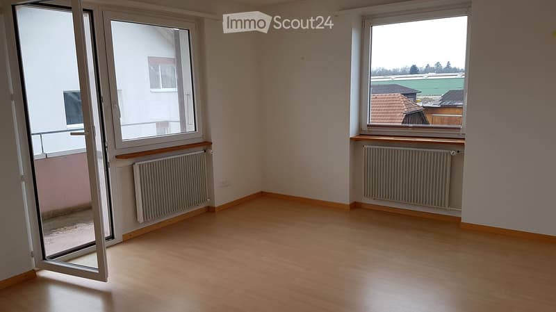 Empty room with wooden flooring, white walls, large windows, and radiators. The room has an open door leading to a balcony or outdoor area.