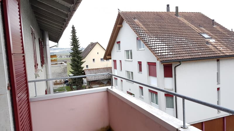Balcony with metal railing overlooking a residential area with houses, trees, and a distant landscape.