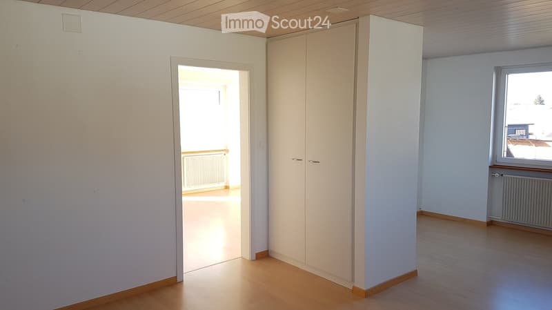 Empty room with light wooden flooring, white walls, and a built-in closet. A doorway leads to another room, and a window offers a view of the outside.