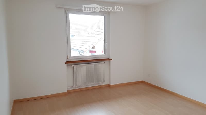 Empty room with wooden flooring, white walls, a window with a view of a roof, and a radiator below the window.