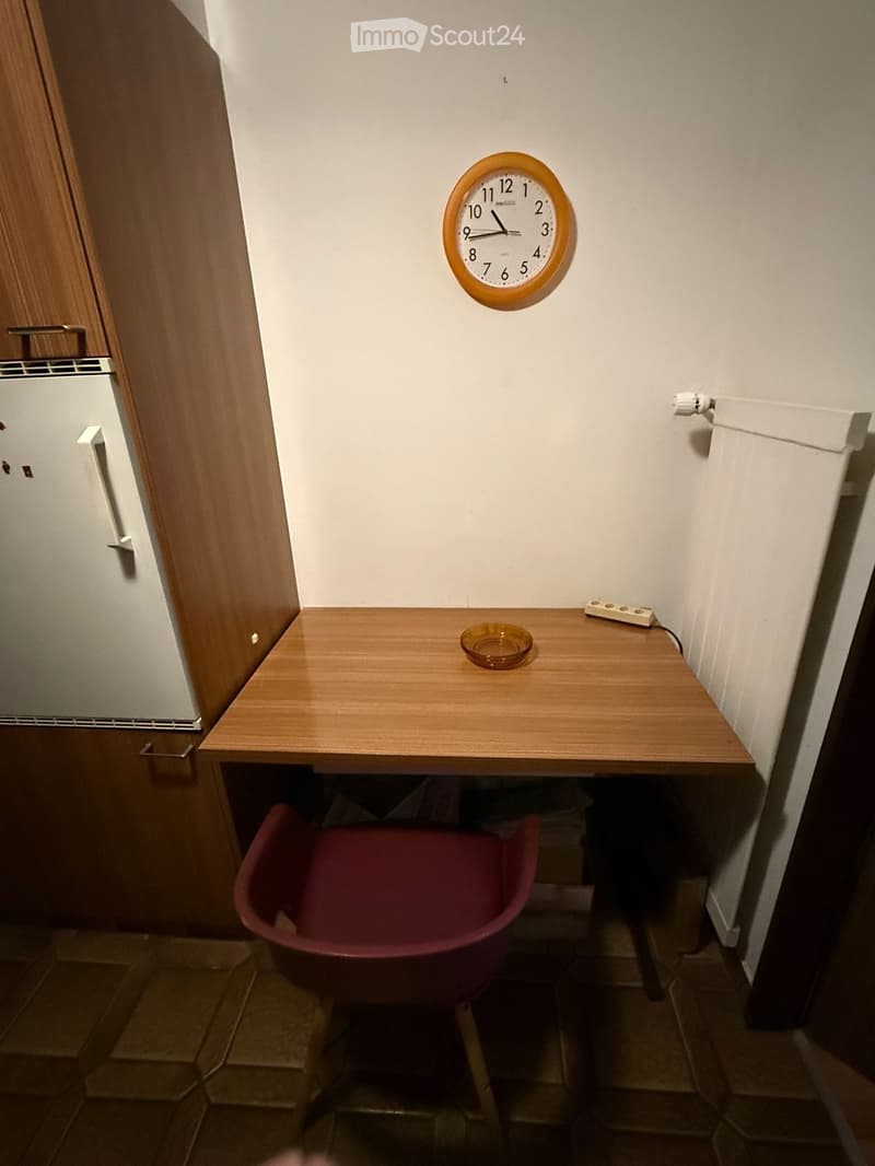 A small, furnished room with a wooden desk, a red chair, a wall clock, and an ashtray on the desk.