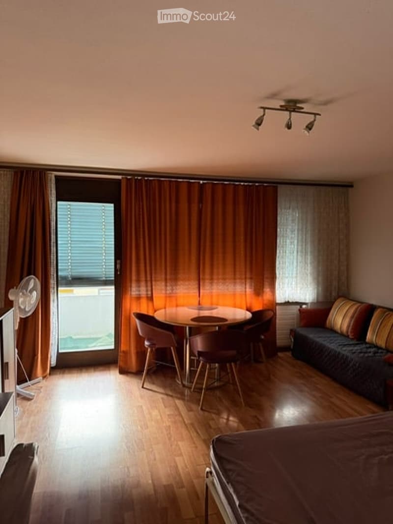 A well-lit room with wooden flooring, a sofa, a dining table with chairs, a bed, and a fan near the window.