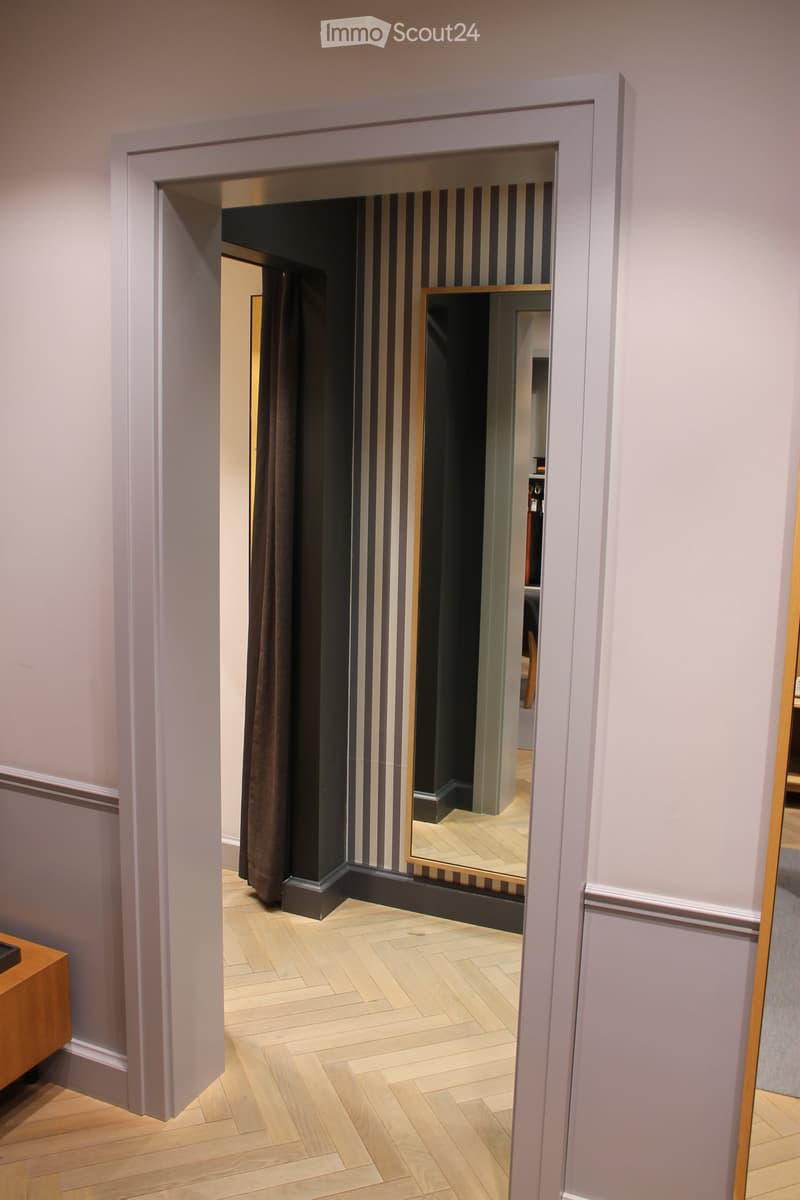A narrow hallway with light wooden flooring, white walls, and a large mirror reflecting part of the adjacent room.