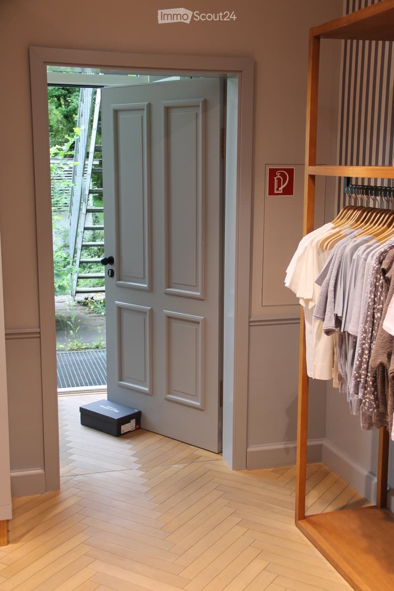 Hallway with wooden herringbone flooring, grey walls, open door leading to garden, clothing rack with hanging garments, and a black box on the floor near the door.
