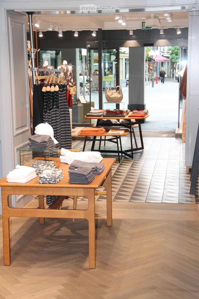 A modern retail store interior with wooden flooring, display tables with folded clothing, a handbag on a shelf, and mannequins dressed in clothing. The space is well-lit with track lighting and has large windows showing the street outside.