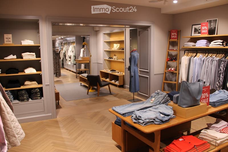 A well-lit clothing store with wooden flooring, shelves displaying folded clothes, a clothing rack with hanging garments, a mannequin in a blue dress, and a table with folded jeans and a handbag.