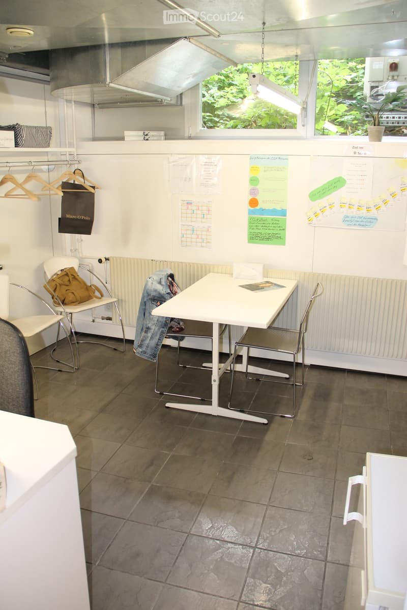 A small, modern room with a white table, two chairs, a coat hanger, and a shelf with a handbag and jeans draped over it. The room has large windows with a view of greenery and is well-lit with natural light.